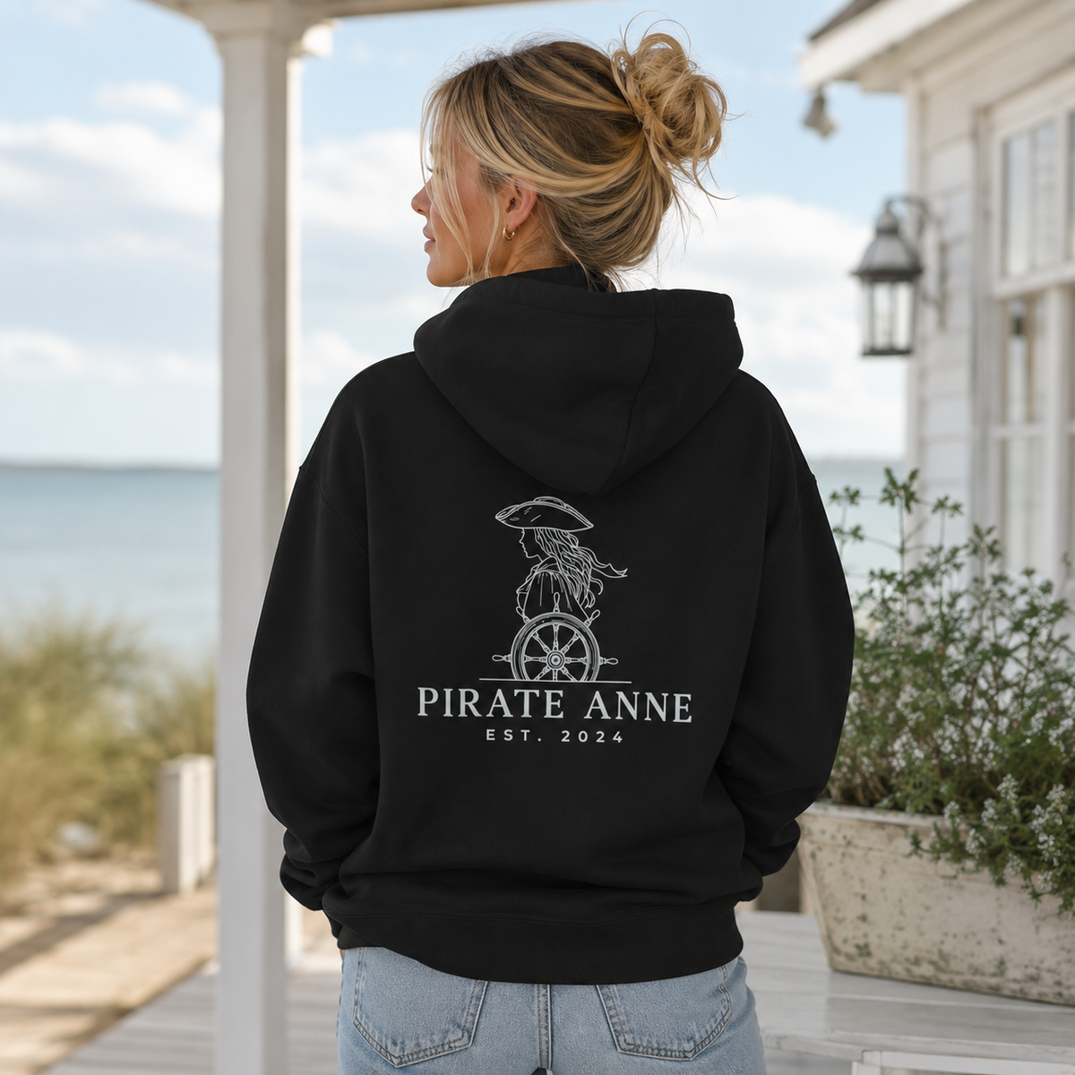 A woman wearing a black hoodie with a white pirate-themed graphic and the text 'PIRATE ANNE EST. 2024' on the back, standing on a porch with the ocean and house in the background.