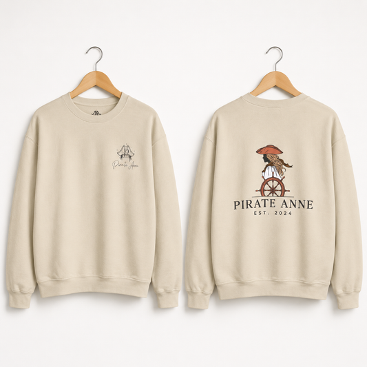 Two cream-colored crewneck sweatshirts on wooden hangers, showing the front and back designs. The front has a small graphic on the left chest, and the back features a larger graphic of a pirate riding a cart with the text 'PIRATE ANNE EST.
