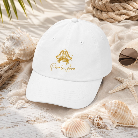 A white baseball cap with gold embroidered text reading 'Pirate Anne' and a gold anchor graphic, styled on a beach-themed surface with seashells, rope, sunglasses, and palm leaves.